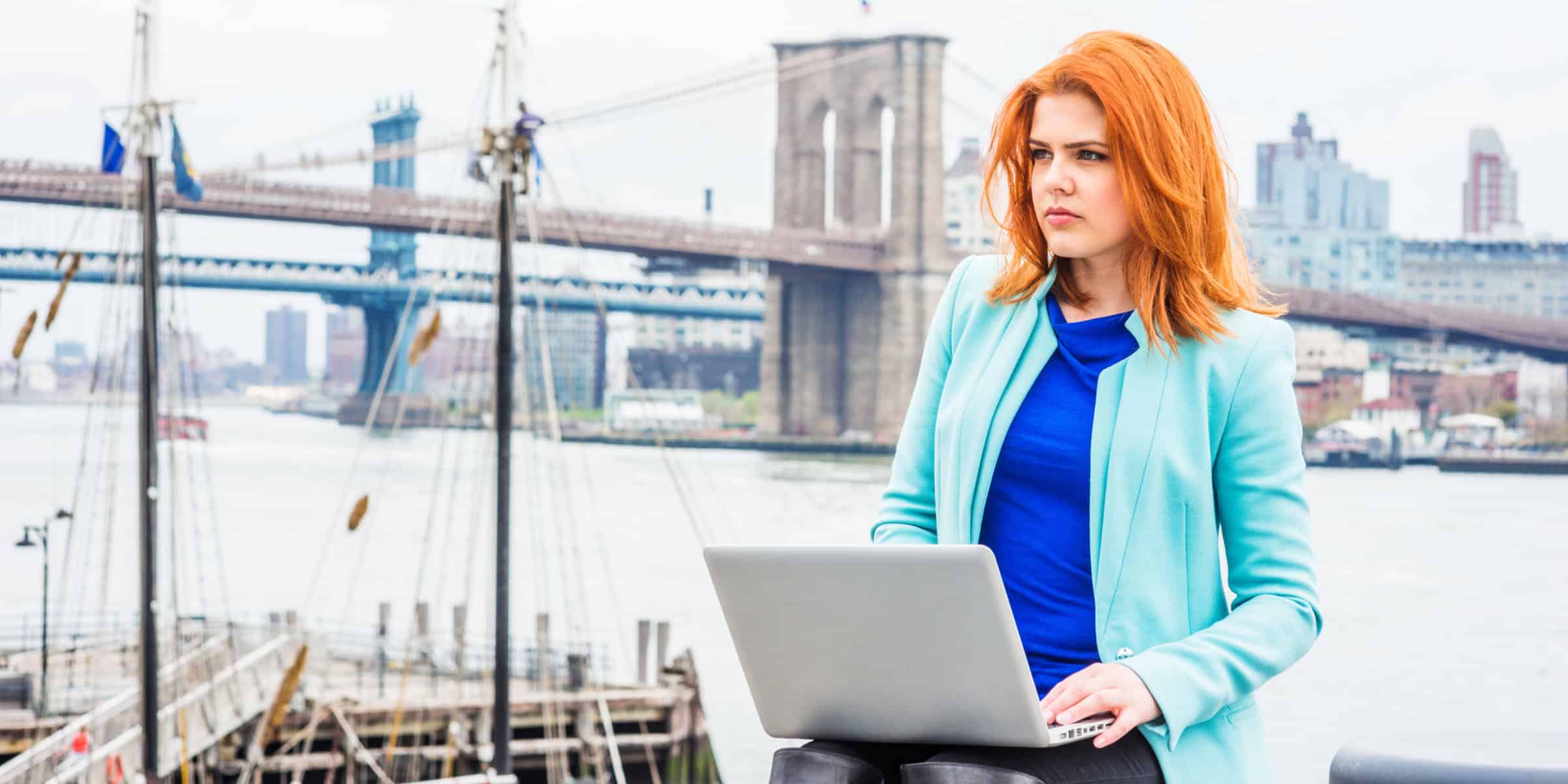 A woman with red hair is using a laptop in front of the brooklyn bridge.