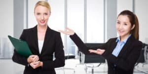 Two women in business suits pointing at each other.