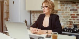 A woman in glasses sitting at a kitchen table with a laptop.