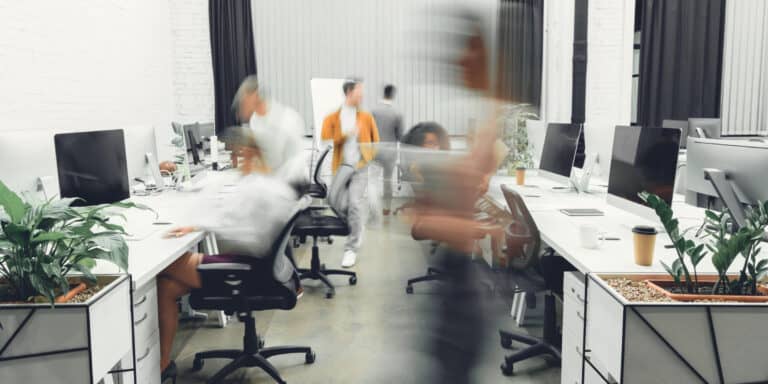 A blurry image of people working at desks in an office.