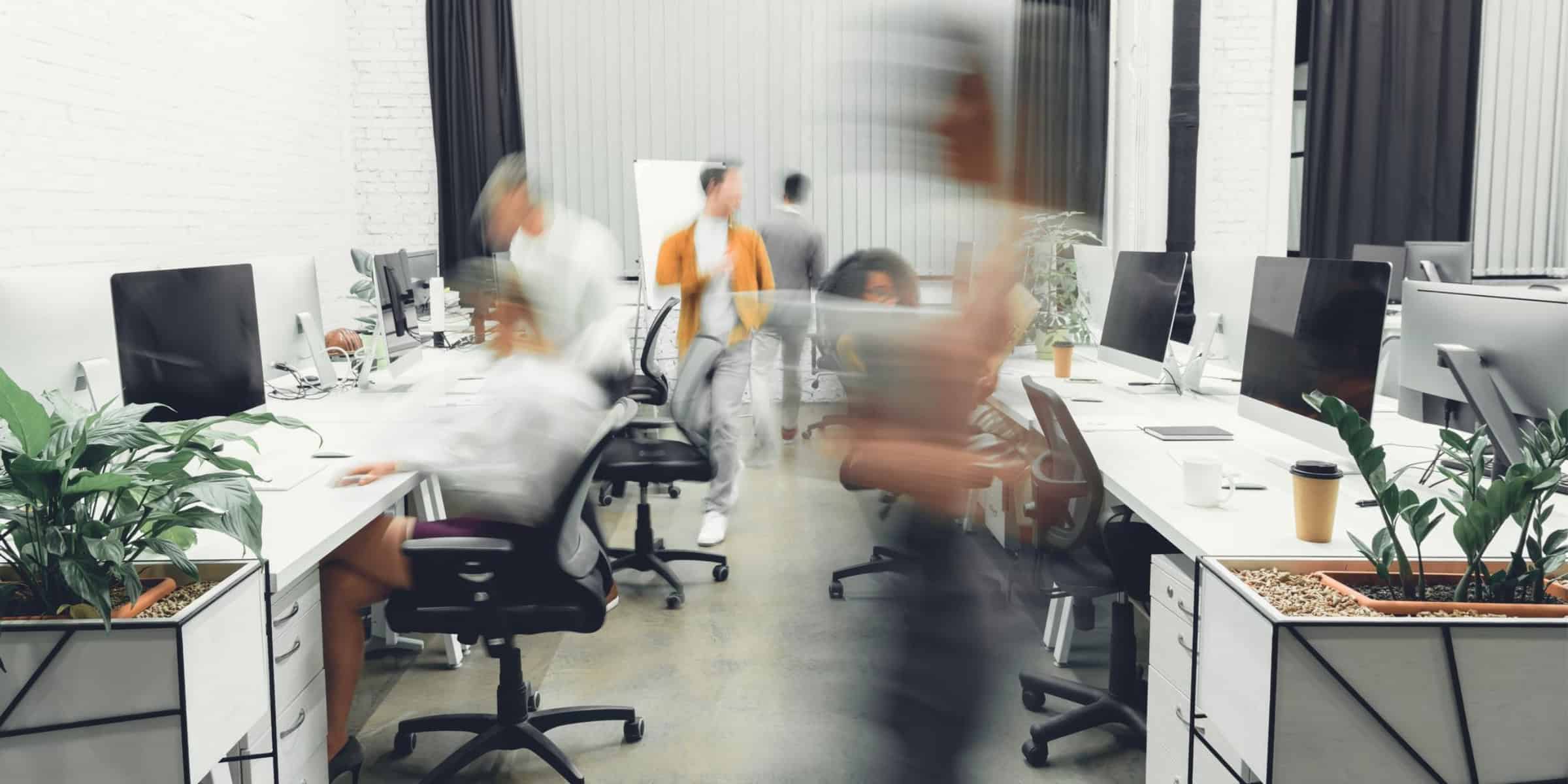 A blurry image of people working at desks in an office.