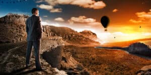 A man is standing on a cliff overlooking a valley with a hot air balloon.