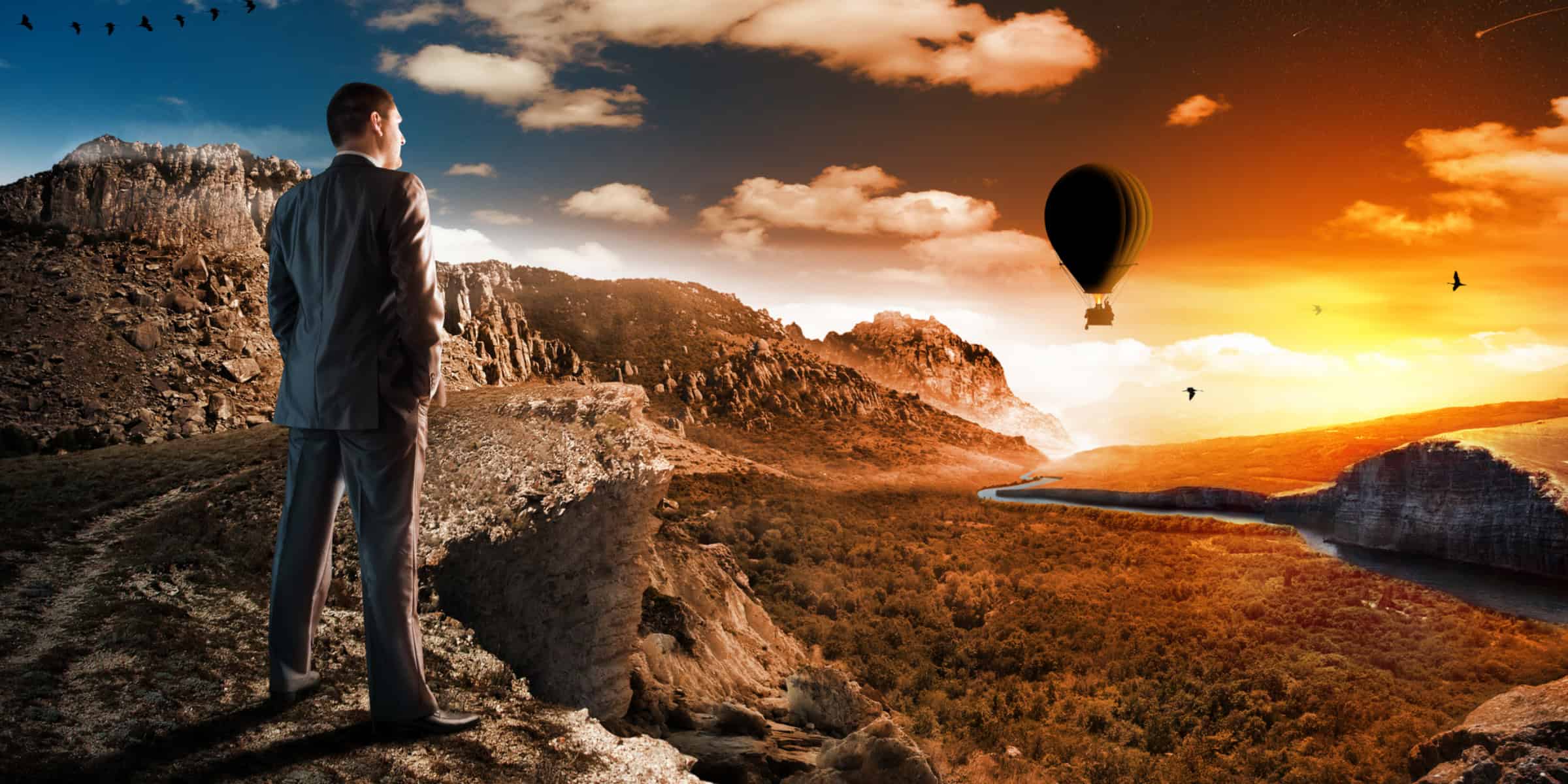 A man is standing on a cliff overlooking a valley with a hot air balloon.