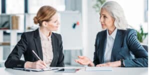 Two professional women in business suits engaged in a discussion at a table in an office setting. one is writing notes.