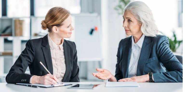 Two professional women in business suits engaged in a discussion at a table in an office setting. one is writing notes.