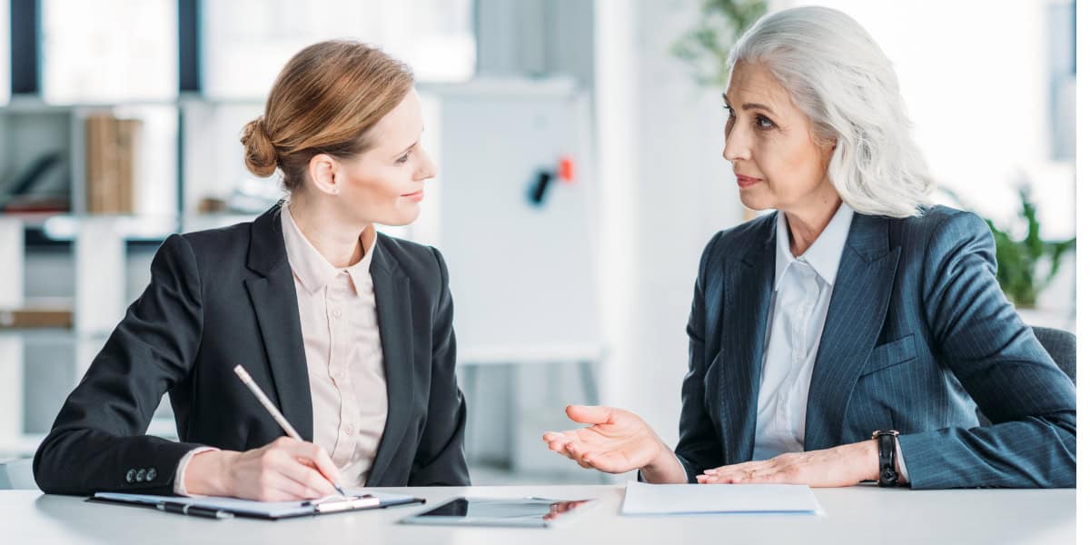 Two professional women in business suits engaged in a discussion at a table in an office setting. one is writing notes.