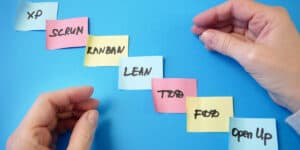 Hands arranging colorful sticky notes labeled with project management terms like "scrum," "kanban," and "lean" on a blue background.