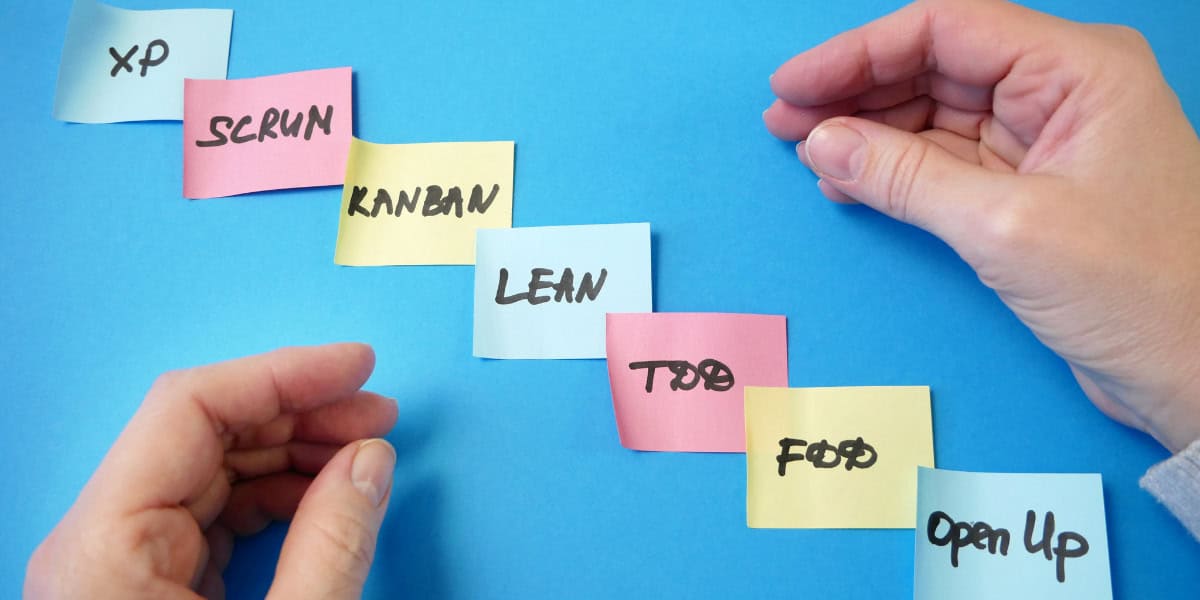 Hands arranging colorful sticky notes labeled with project management terms like "scrum," "kanban," and "lean" on a blue background.