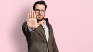 A man wearing glasses and a suit making a stop sign on a pink background.
