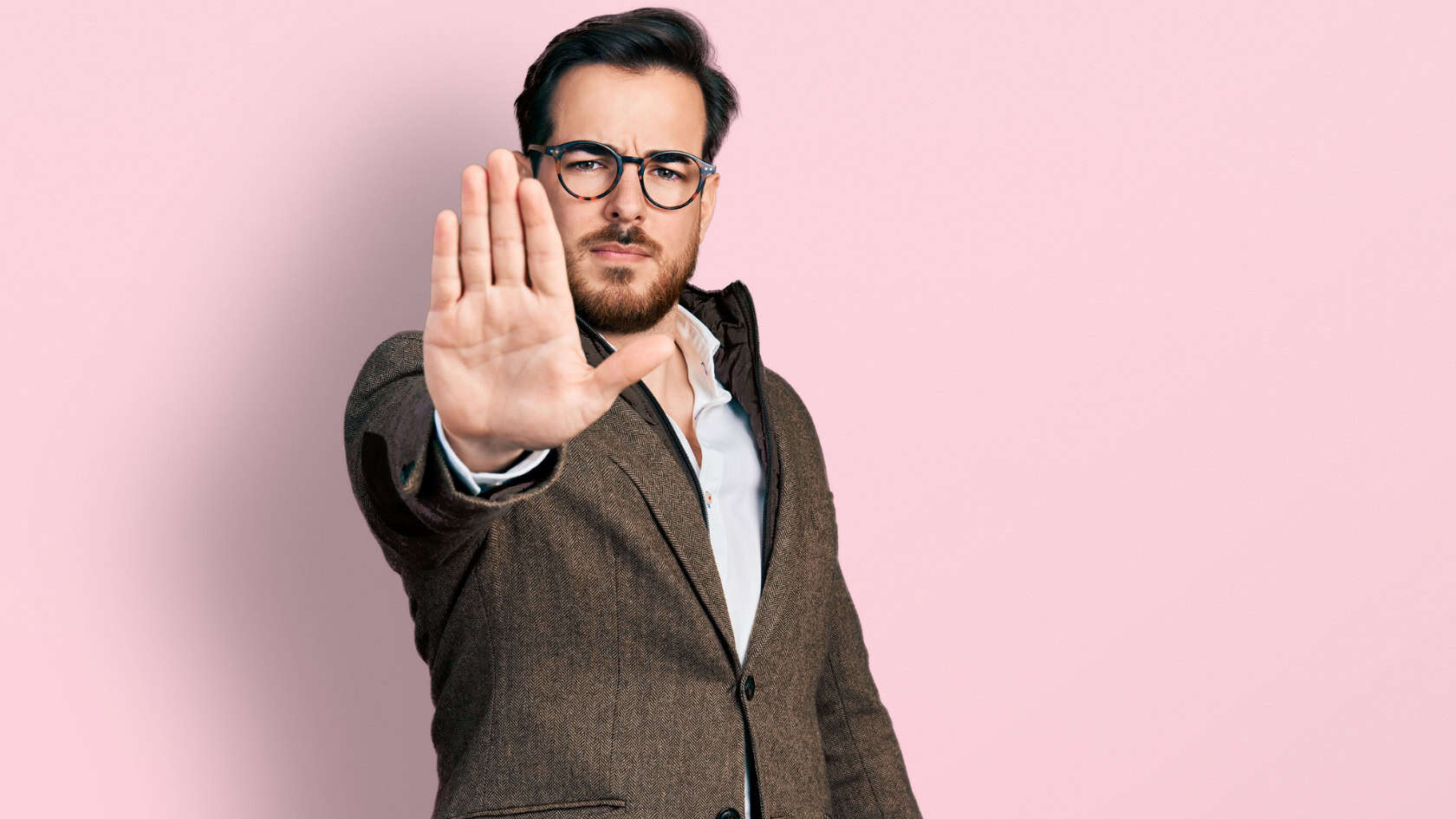 A man wearing glasses and a suit making a stop sign on a pink background.