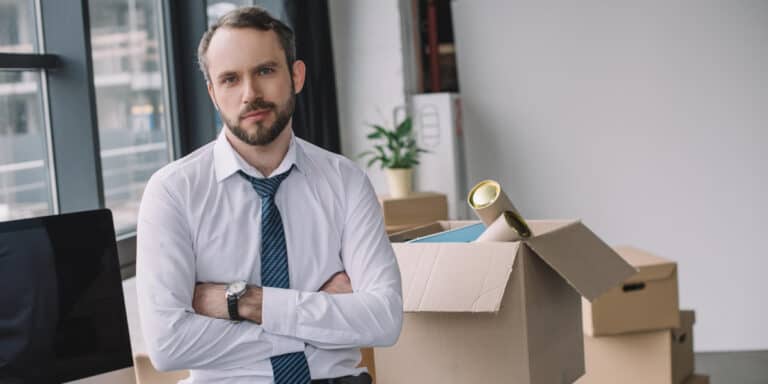A man in a white shirt and tie standing with arms crossed in an office, with cardboard boxes and a plant in the background.