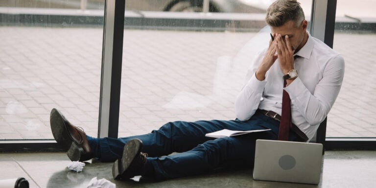 Businessman sitting on the floor looking stressed with documents and a laptop nearby.