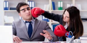 A woman wearing boxing gloves pretends to punch a man in a suit and glasses in an office setting. The man appears surprised or startled. Bookshelves and office supplies are visible in the background.