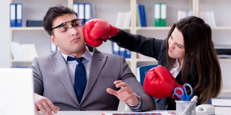 A woman wearing boxing gloves pretends to punch a man in a suit and glasses in an office setting. The man appears surprised or startled. Bookshelves and office supplies are visible in the background.
