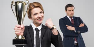 A woman in a business suit holds a trophy and smiles while a man in a business suit stands with crossed arms in the background.
