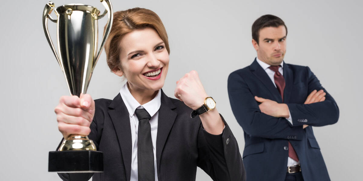 A woman in a business suit holds a trophy and smiles while a man in a business suit stands with crossed arms in the background.