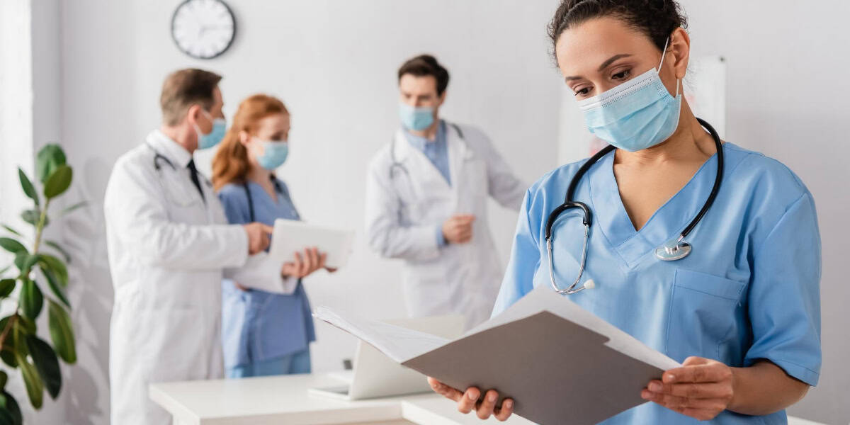 Healthcare professionals wearing masks and scrubs in a medical office; a female nurse in the foreground reads a file while three other professionals converse in the background.