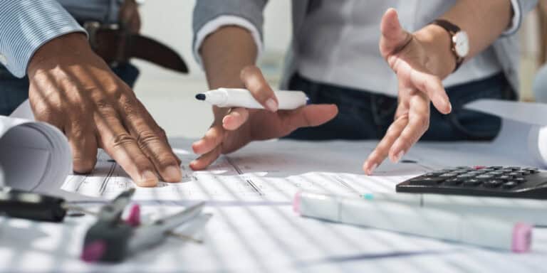 Two people discuss a project over architectural blueprints on a table, with a calculator, pen, and drafting tools visible nearby.