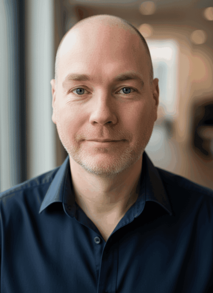 A bald man with a trimmed beard, wearing a navy blue shirt, stands indoors near a window with a softly blurred background.